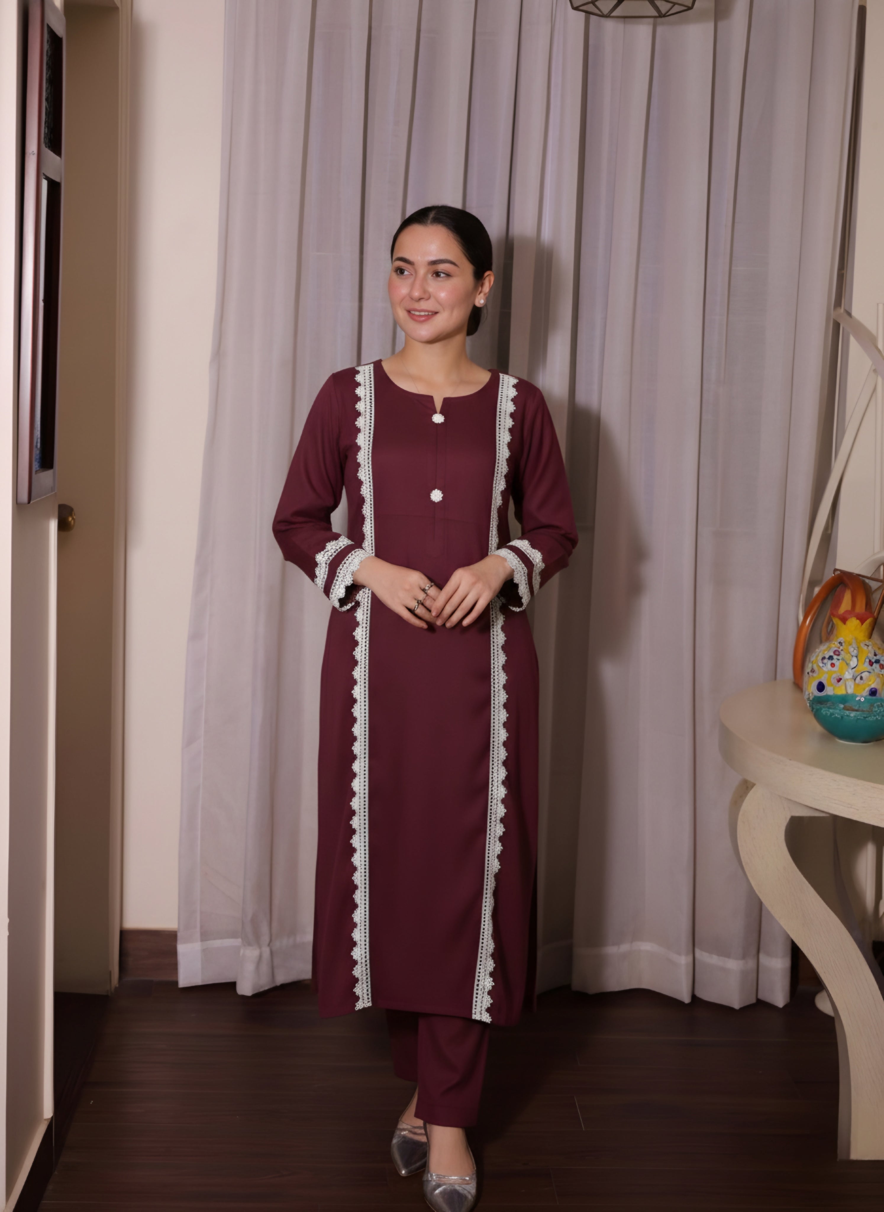 Woman wearing a maroon traditional outfit with white trim standing indoors against a curtain.